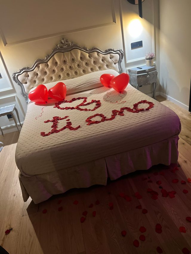 Letto decorato con petali di rosa a forma di cuore, scritta «ti amo» e palloncini